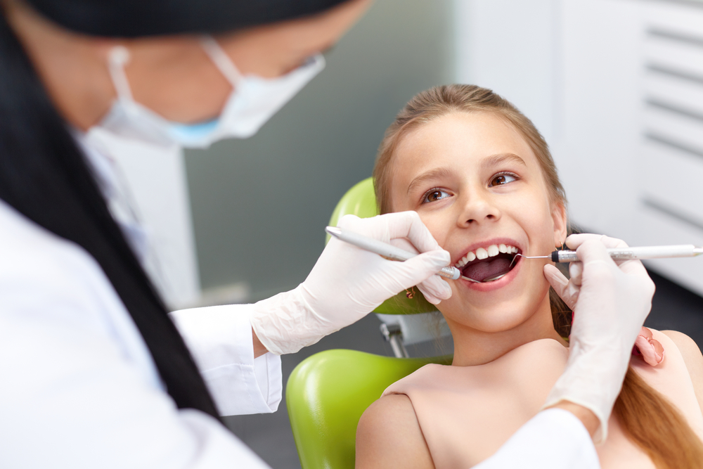 A young girl smiling in a dental chair while a dentist in a mask examines her teeth with dental instruments - Dentist University Place WA A young girl smiling in a dental chair while a dentist in a mask examines her teeth with dental instruments - Dentist University Place WA