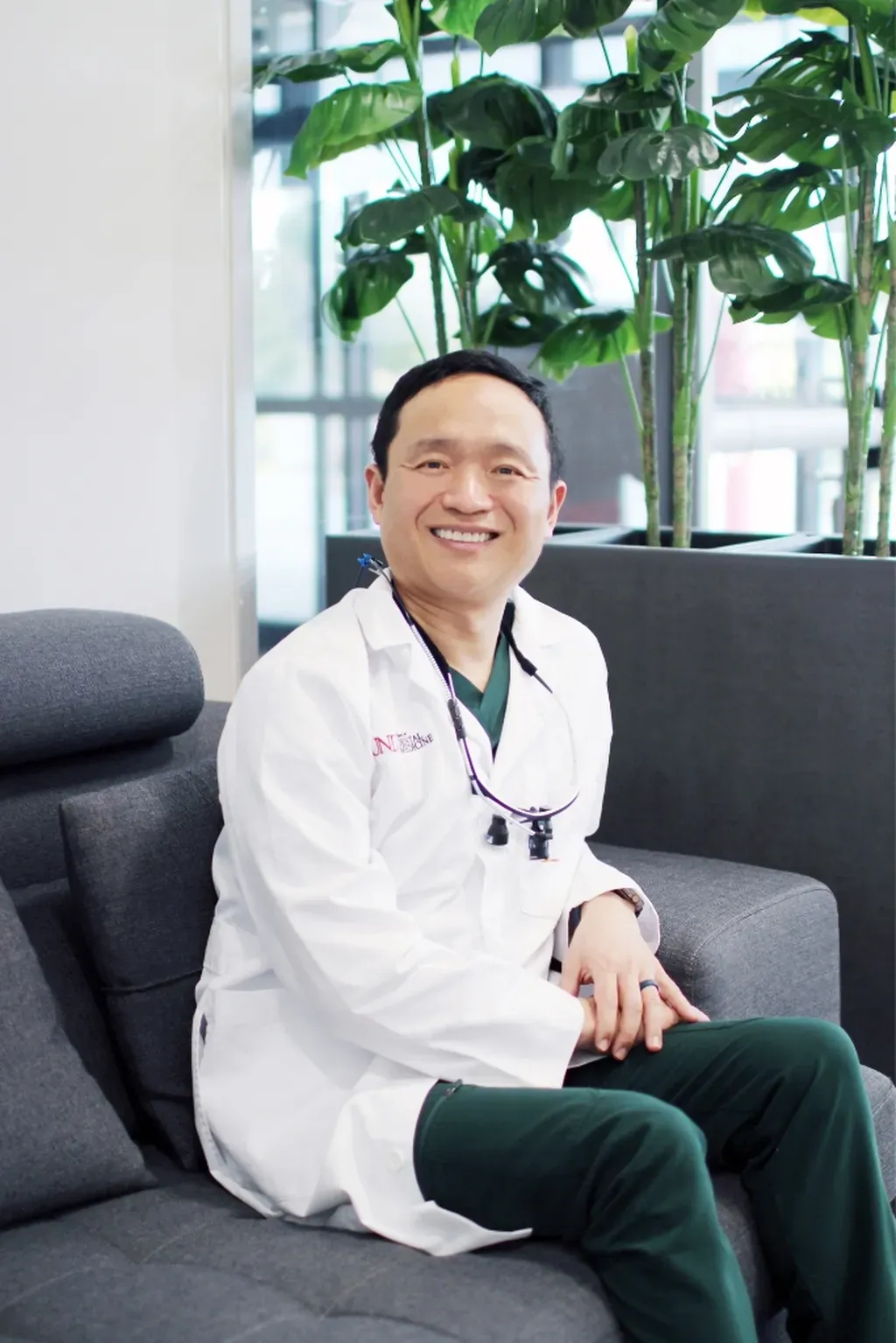 Dr. Hanbit Chi, DDS in a white coat and green scrubs sits on a gray chair, smiling, with tall green plants in the background - Dentist University Place WA Dr. Hanbit Chi, DDS in a white coat and green scrubs sits on a gray chair, smiling, with tall green plants in the background - Dentist University Place WA