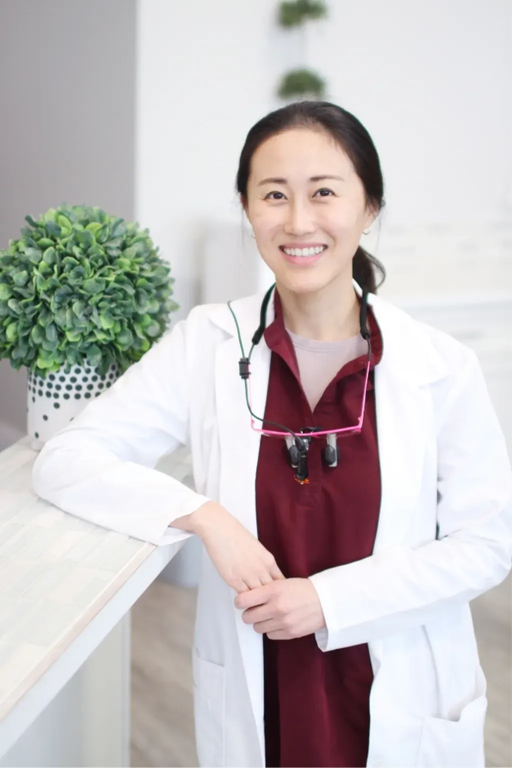 Dr. Junkyung Lee, DMD in a white coat and burgundy scrubs smiles while standing near a countertop with a small green plant - Dentist University Place WA Dr. Junkyung Lee, DMD in a white coat and burgundy scrubs smiles while standing near a countertop with a small green plant - Dentist University Place WA