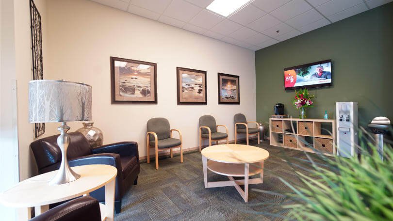 A cozy dental office waiting room with chairs, a TV, framed artwork, and a water cooler, decorated with flowers and a modern lamp - Dentist University Place WA A cozy dental office waiting room with chairs, a TV, framed artwork, and a water cooler, decorated with flowers and a modern lamp - Dentist University Place WA